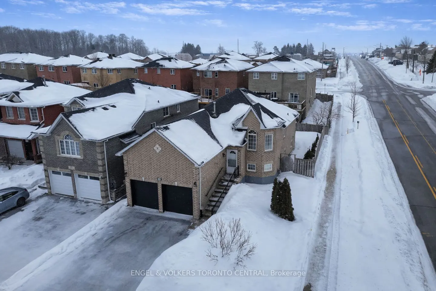 Aerial view of a two-story brick house with a black roof covered in snow, a two-car garage, and a snow-covered driveway. Other houses and a road are visible.