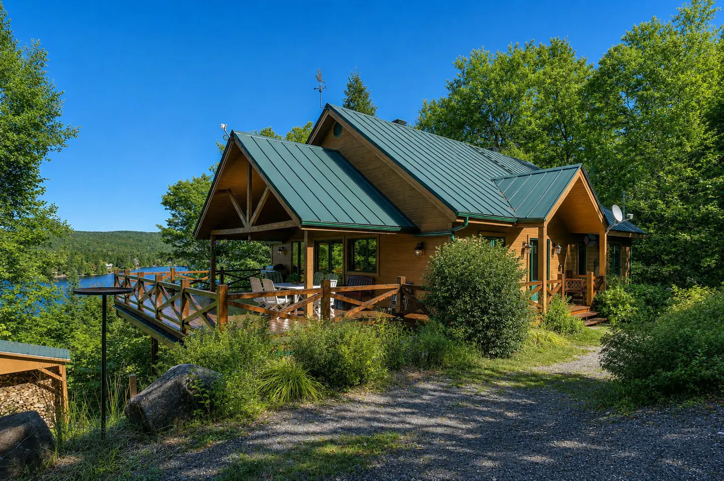 A wooden cabin with a green metal roof, surrounded by trees, overlooks a lake on a sunny day.