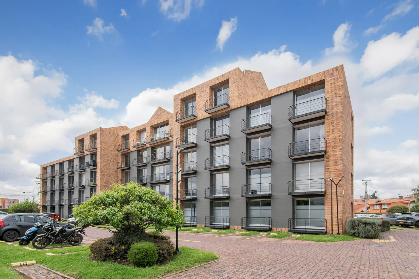 Modern apartment building with gray walls, brick accents, and black balconies under a blue sky. Cars and greenery in the foreground.