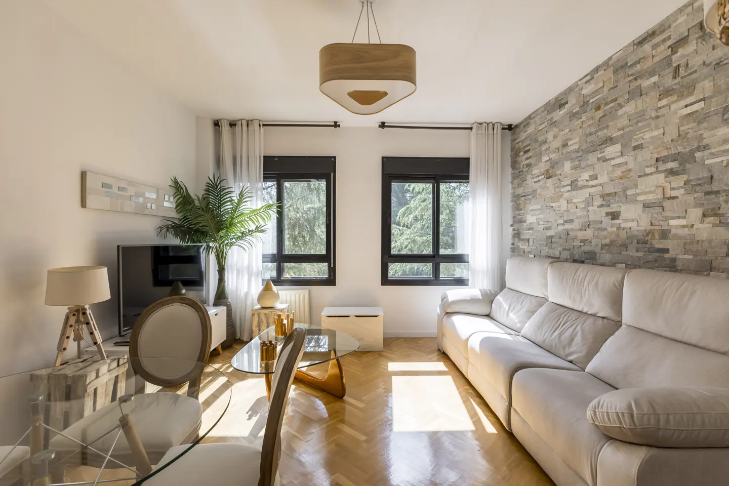 Living room with a stone accent wall, a beige sofa, and a glass dining table with wooden chairs. Two windows with black frames let in natural light.