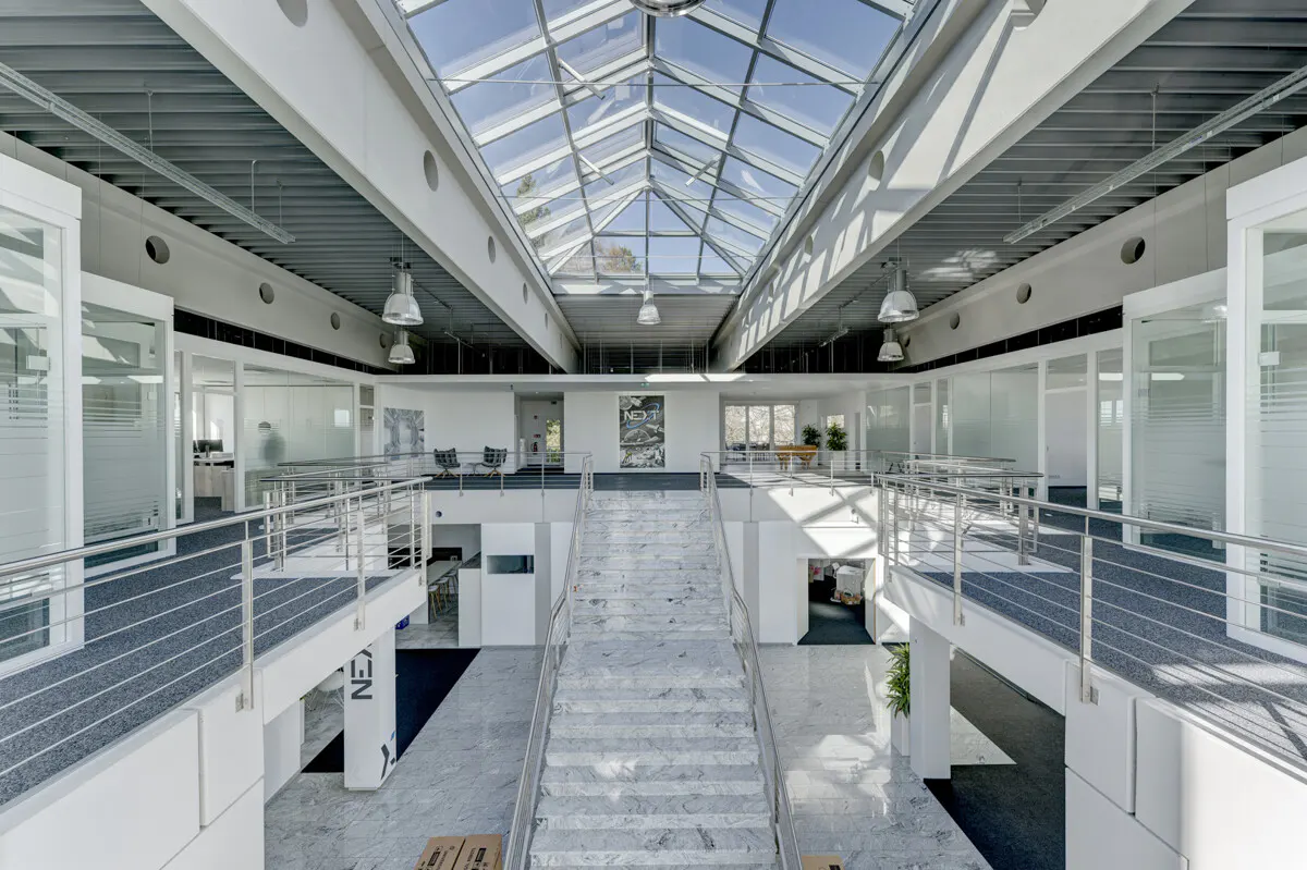 Bright, modern office interior with a marble staircase, glass roof, and white walls. Balconies with metal railings overlook the open space.