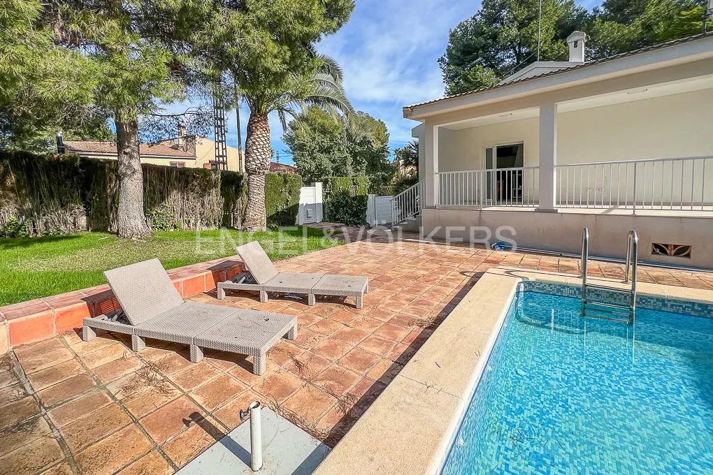 Backyard view with a pool, two lounge chairs on a brick patio, and a white house with a covered porch.