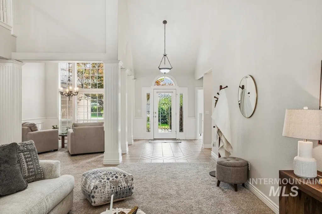Bright, airy foyer with white walls, columns, and a glass front door. Living room with gray furniture is visible to the left.