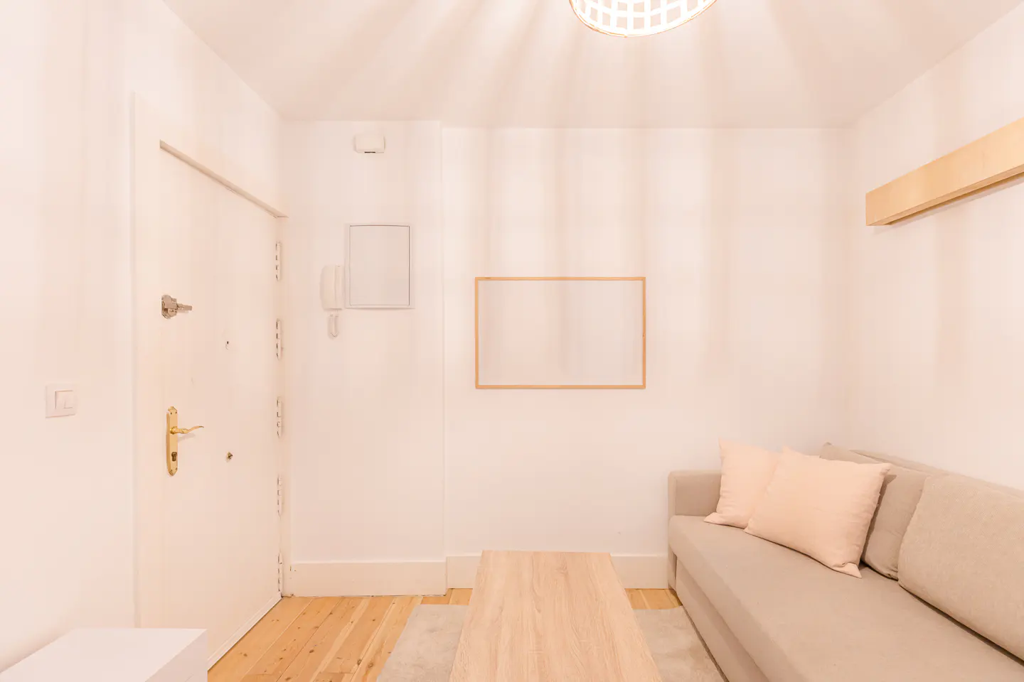 A bright living room with a beige sofa, light wood coffee table, and white walls. A white door is on the left.