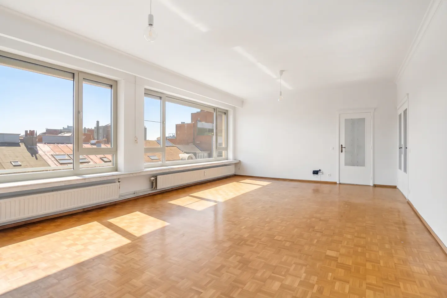 Bright, empty room with parquet floors, white walls, and large windows showing a city view. Radiators line the wall under the windows.