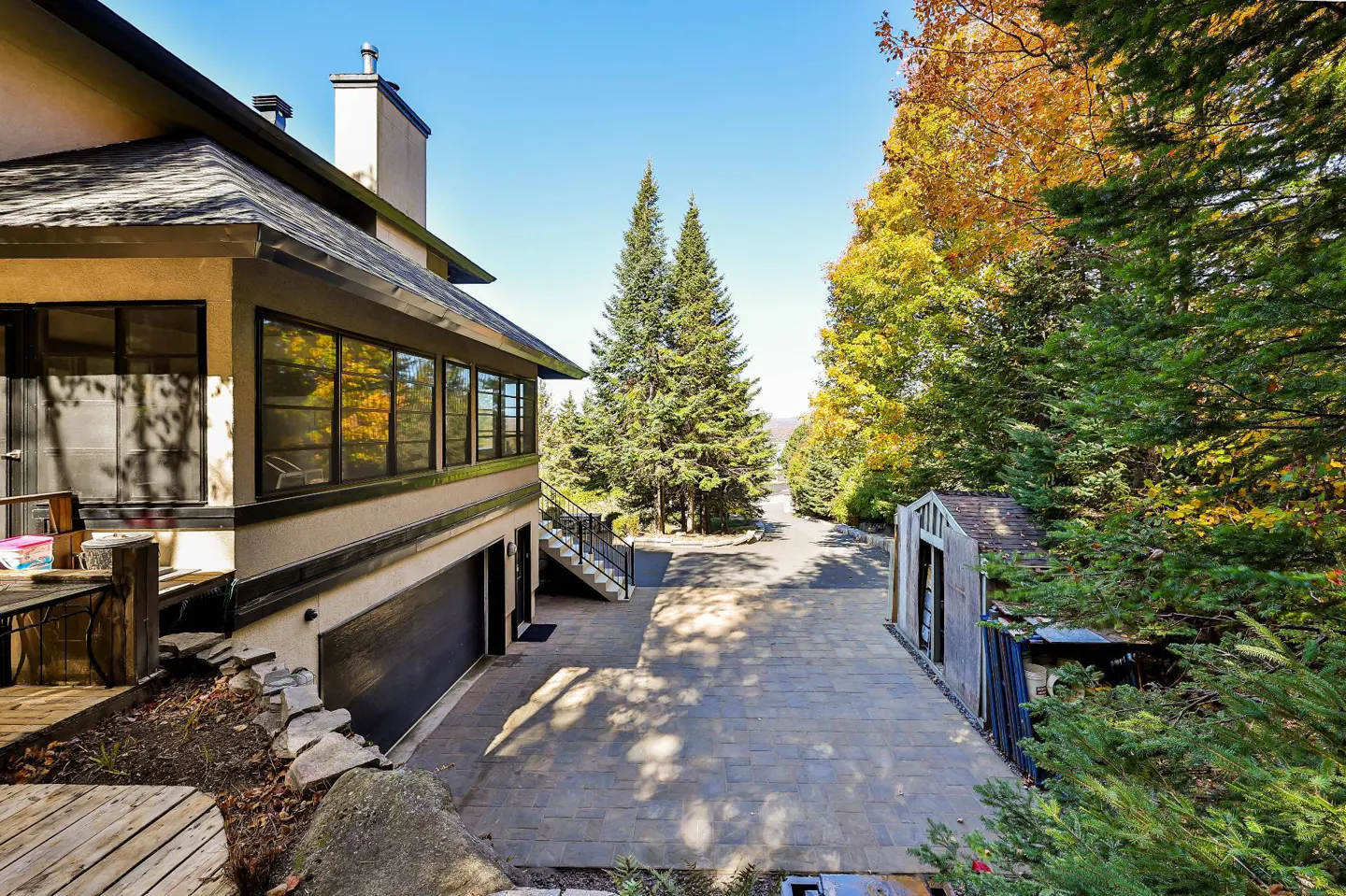 Exterior view of a beige house with a black roof, a paved driveway, and green trees under a blue sky.
