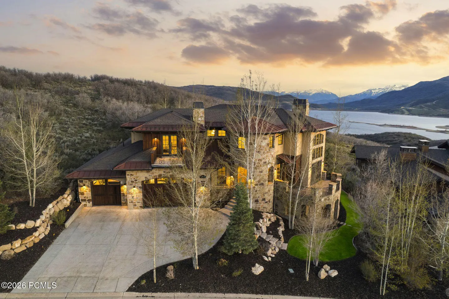 Aerial view of a stone house with a dark roof, a driveway, and a lake and mountains in the background at sunset.
