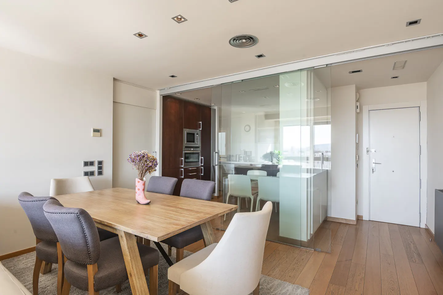 Bright dining area with a wooden table, gray and white chairs, and a pink vase with flowers. A glass wall separates the kitchen.