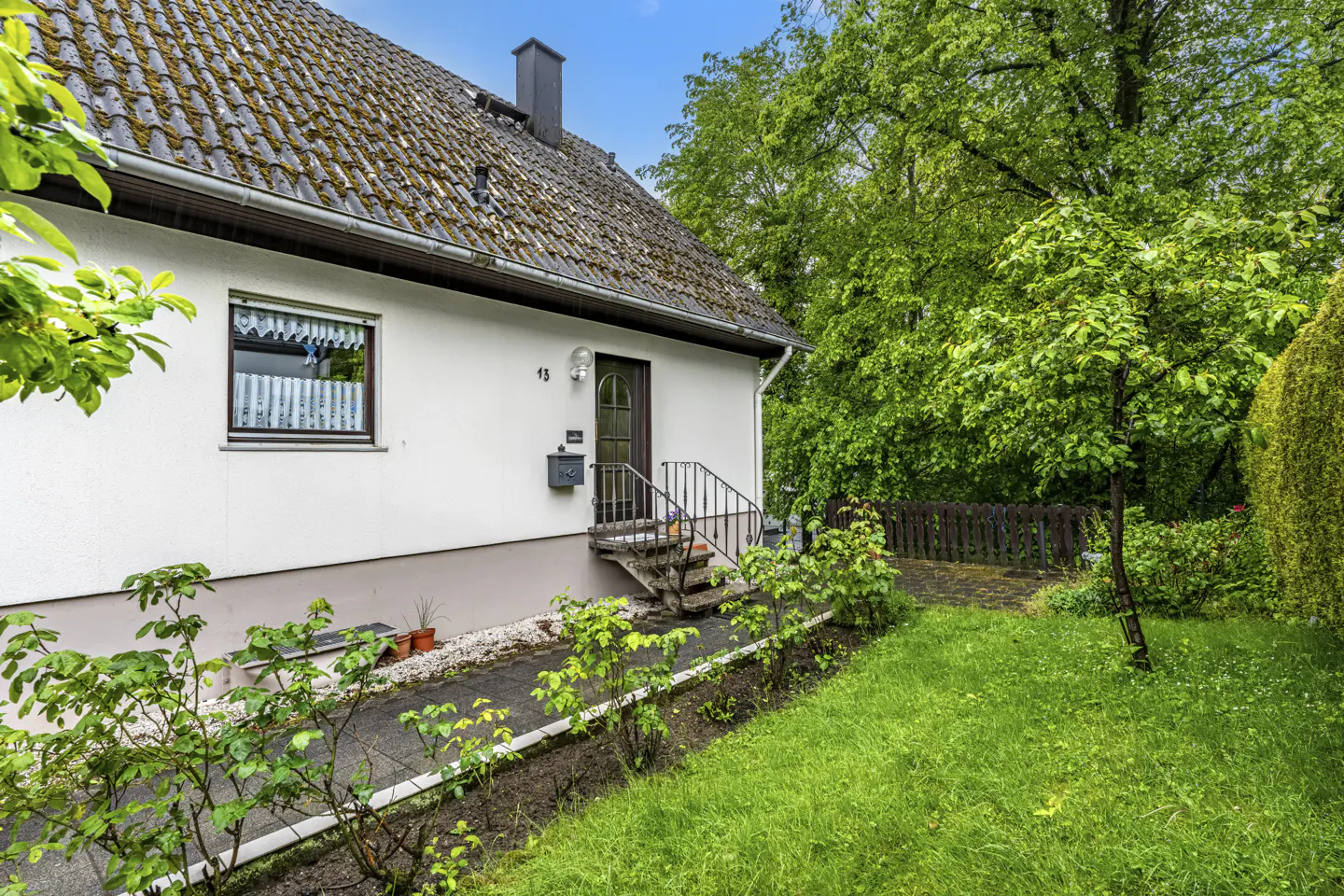 White house with a tiled roof, front entrance with steps, and a lush green garden with trees and a wooden fence.