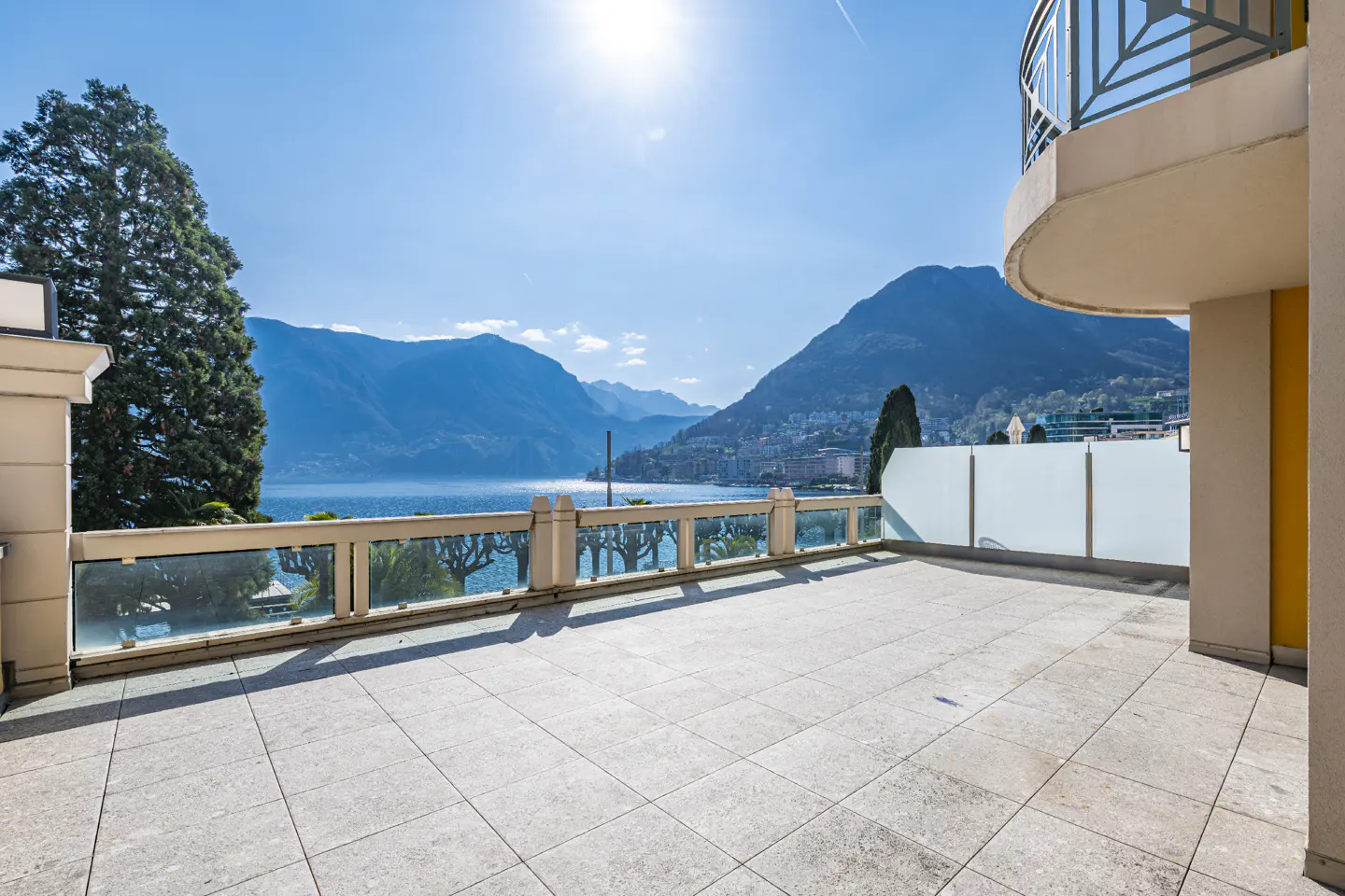 Balcony view of a lake and mountains. The balcony has a stone floor and glass railings. The sky is blue and sunny.
