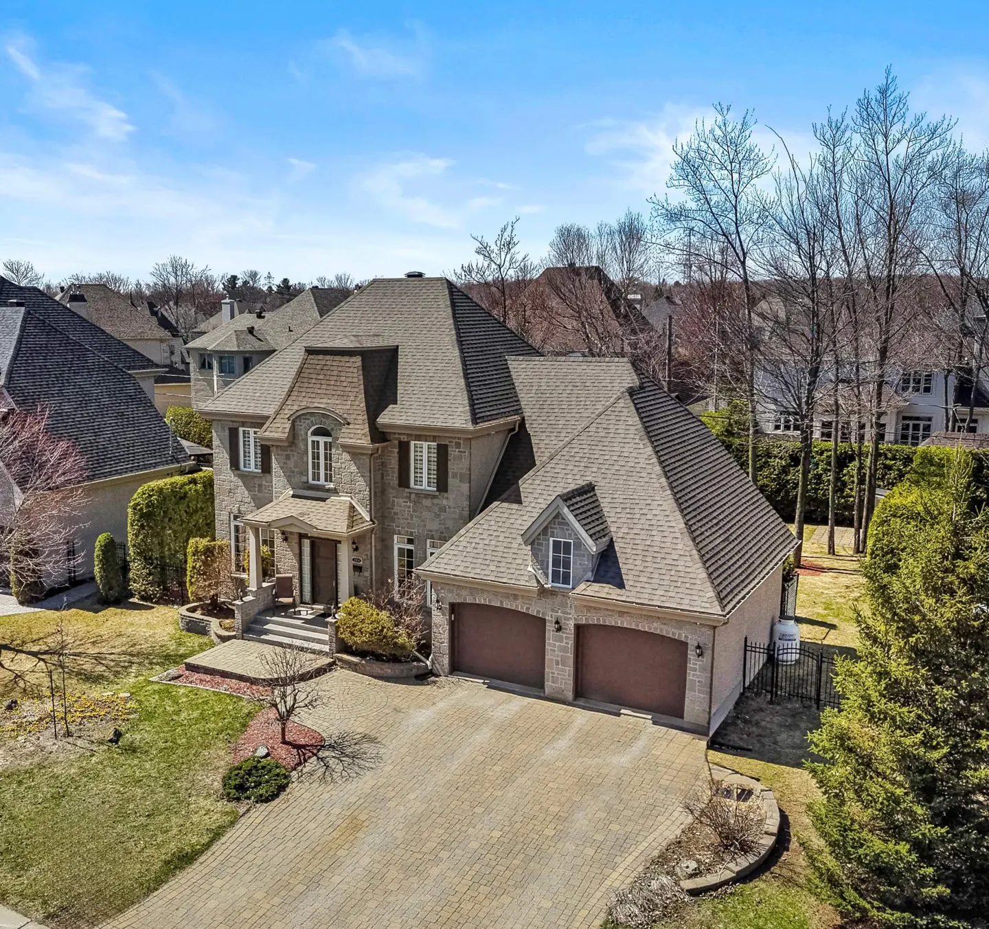 Aerial view of a large, two-story stone house with a brown roof and a two-car garage on a paved driveway.