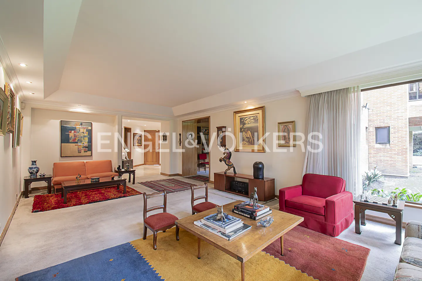 A living room with a red sofa, a wooden table, and a colorful rug. Artwork hangs on the walls.
