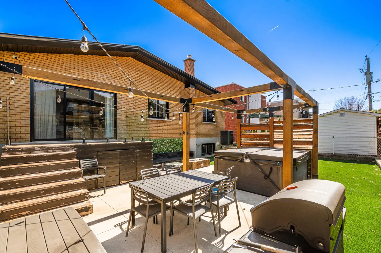 A backyard patio with a table, chairs, grill, and hot tub under a wooden pergola, with string lights and a brick house.