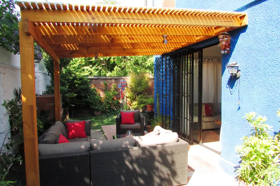 Outdoor patio with a wooden pergola, gray wicker furniture with red pillows, and a blue stucco wall with sliding glass doors.
