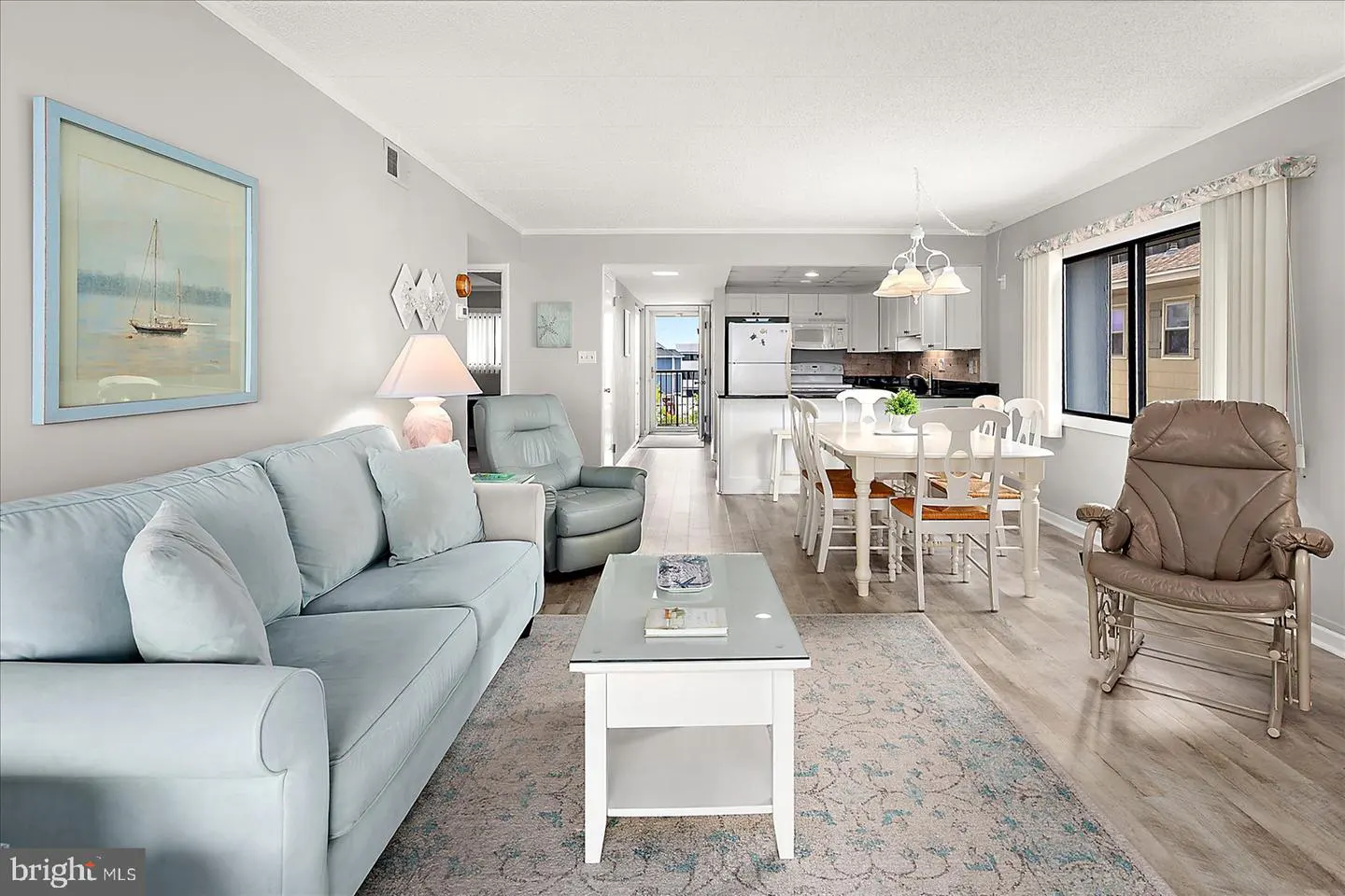 Bright, open-concept living space with a light blue sofa, white dining set, and a view of the ocean through an open doorway.