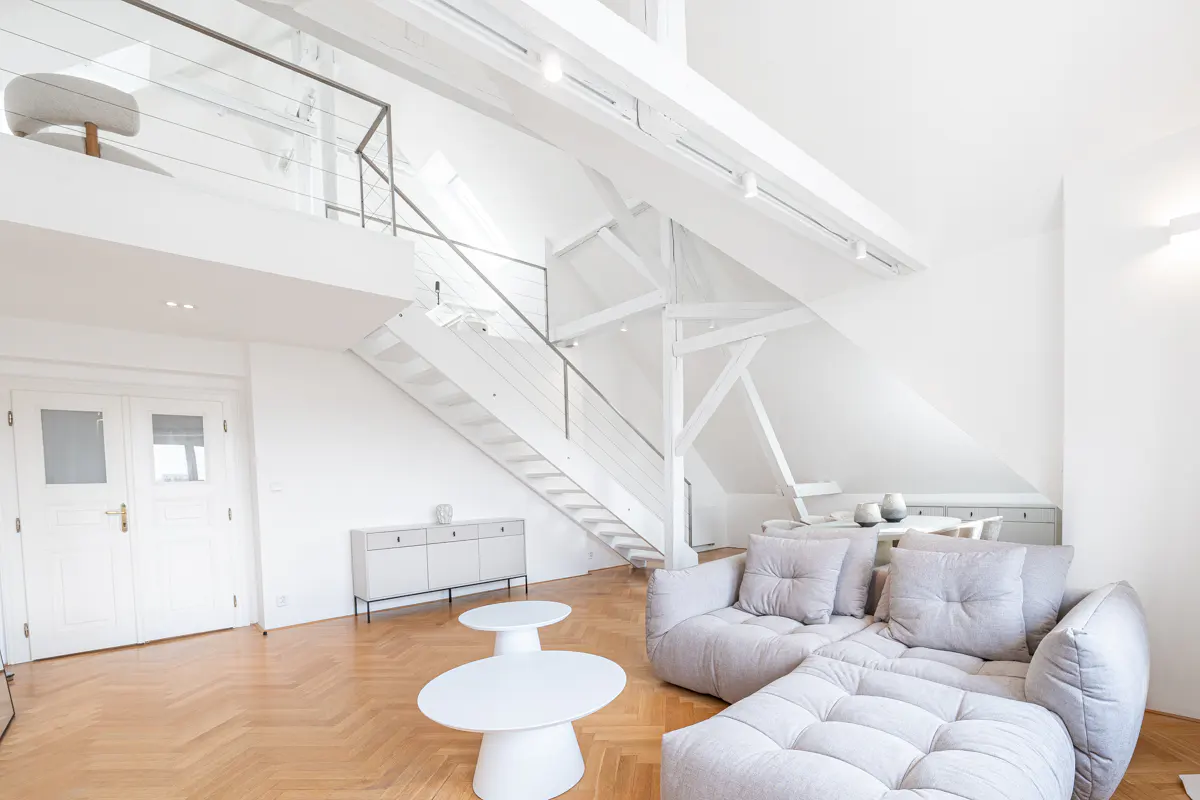 Bright, modern living room with white walls, wood floors, and a gray sectional sofa. A white staircase leads to a loft area.