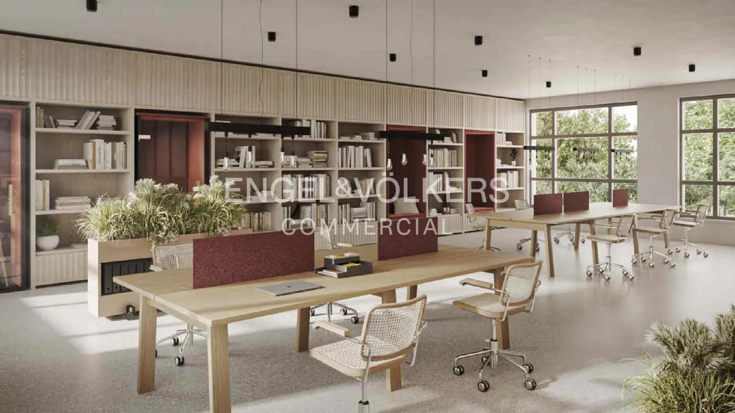 Office space with long wooden tables, rolling chairs, and bookshelves filled with books. Large windows let in natural light.