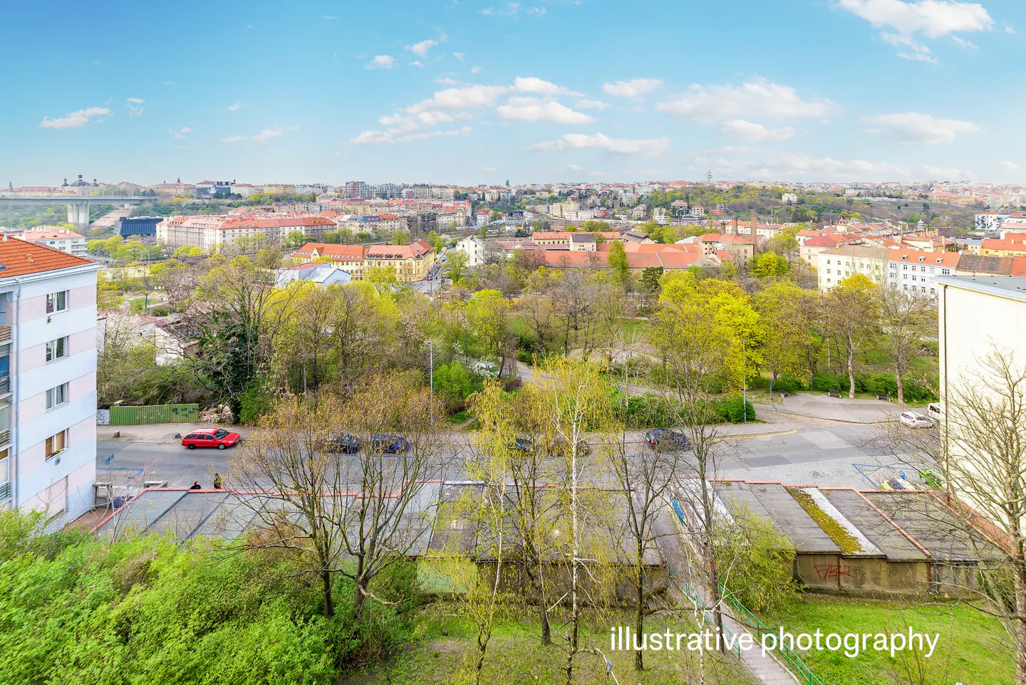 Cityscape view with buildings, trees, and a blue sky with clouds. A red car is parked on the street.