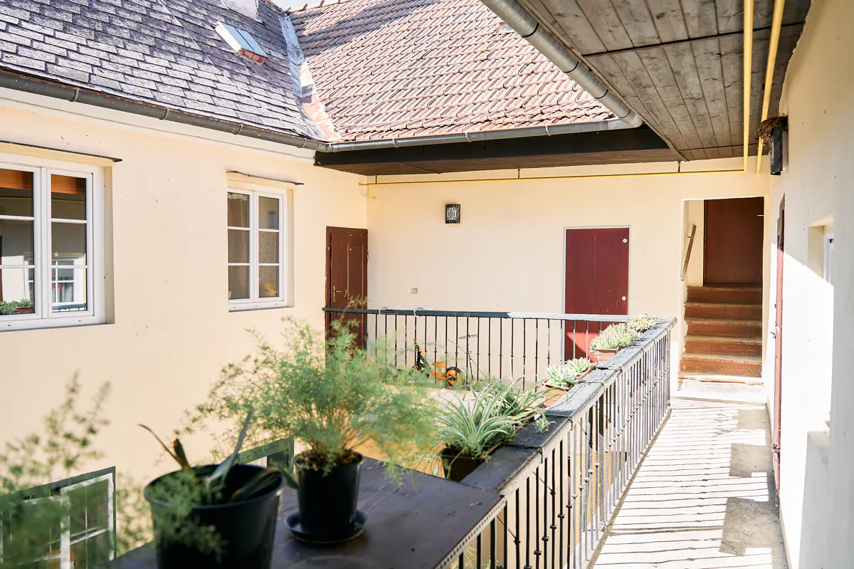 Courtyard view with potted plants on a black railing, cream walls, and brown doors. A tiled roof is visible above.