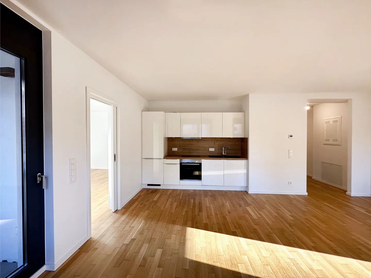 Modern open-plan kitchen with white cabinets, wood backsplash, and light hardwood floors in a bright, minimalist apartment.