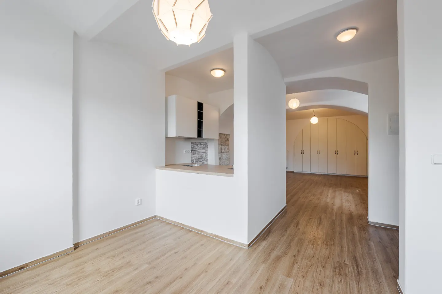 Bright, modern apartment interior with white walls, wood floors, and an open kitchen area. Arched doorway leads to a room with built-in wardrobes.