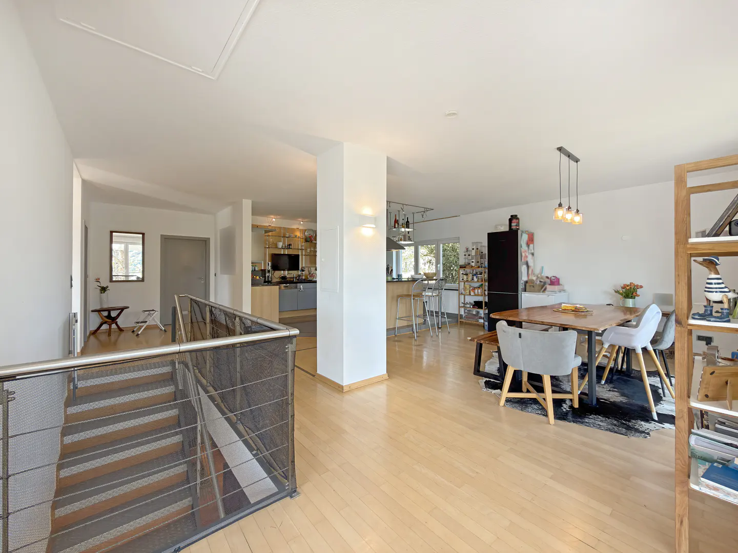 Open-concept home interior with light wood floors, stairs with metal railing, dining table, and kitchen in the background.