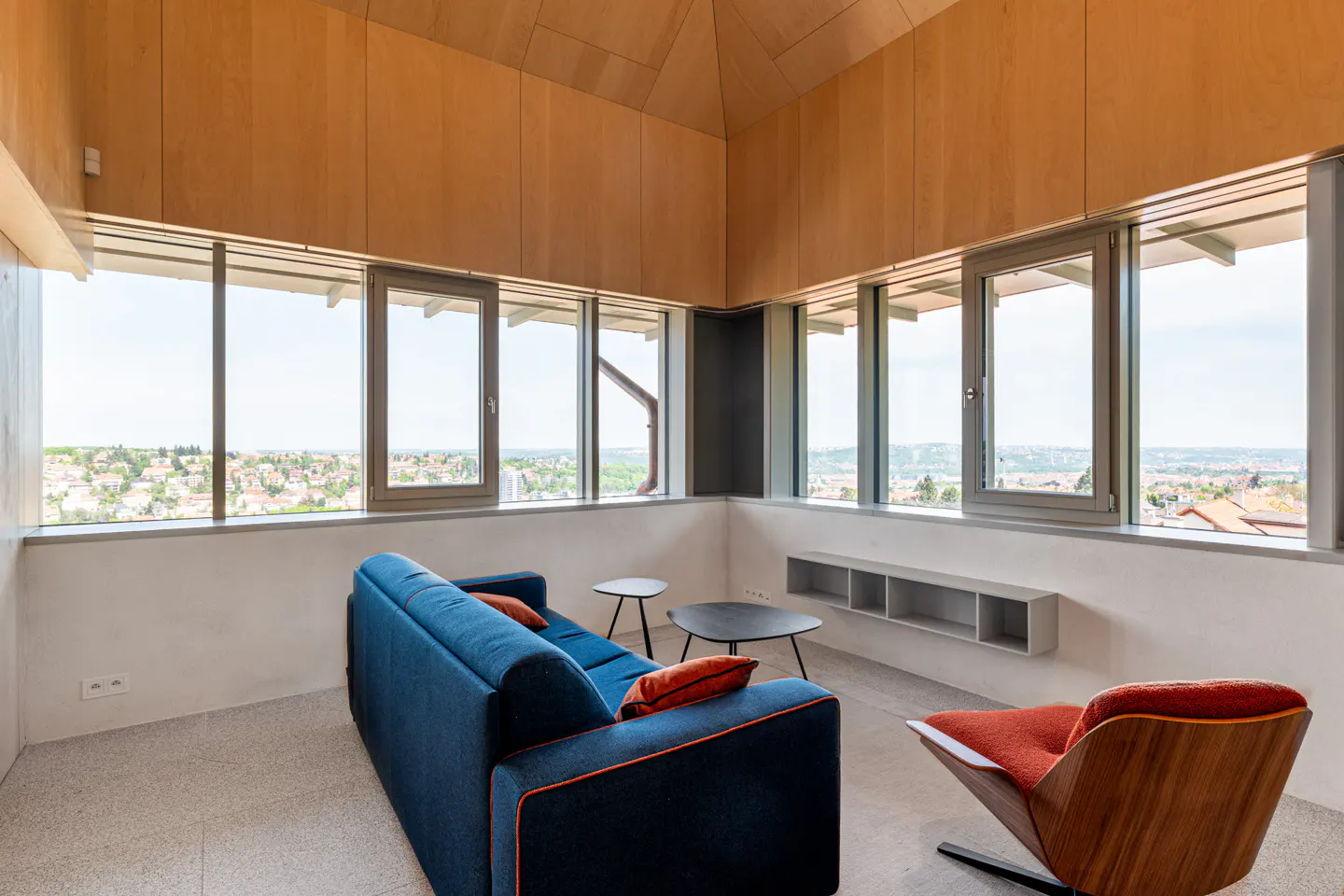 Bright living room with wood paneled walls, large windows, blue sofa, orange chair, and city view.