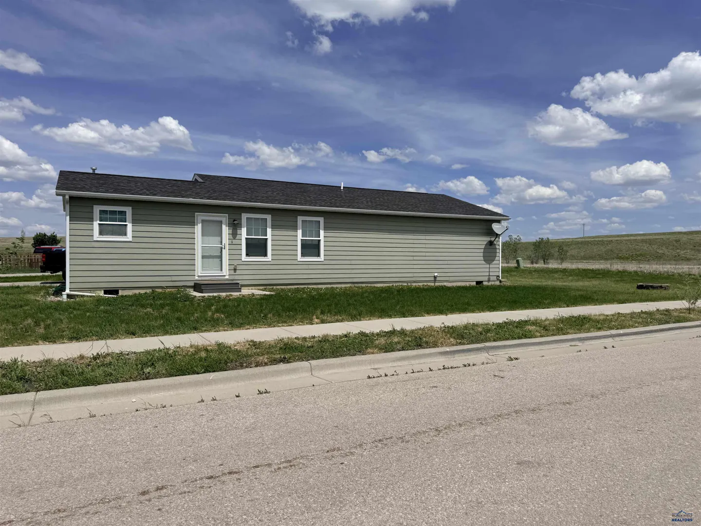 Single-story sage green house with a dark roof, white trim, and a grassy yard under a blue sky with scattered clouds.