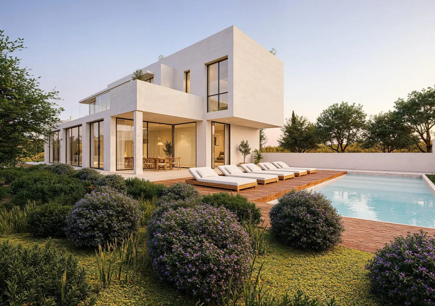 Modern white house with a pool and wooden deck. Lounge chairs line the pool. Green bushes and trees surround the property.