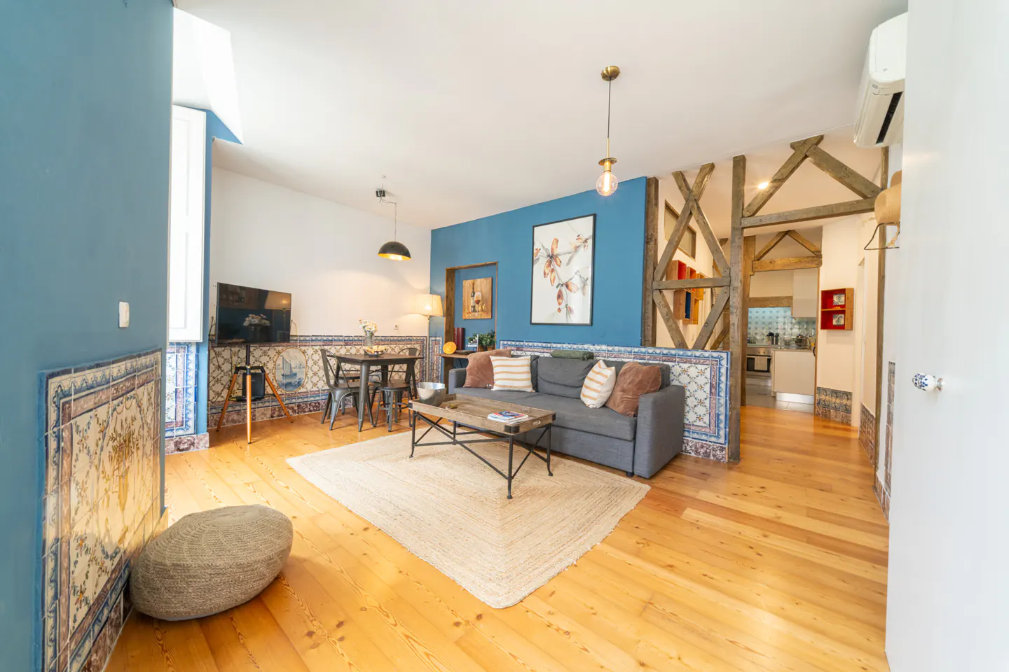 Bright living room with blue walls, wood floors, and tiled accents. A gray sofa, wood coffee table, and dining set furnish the space.