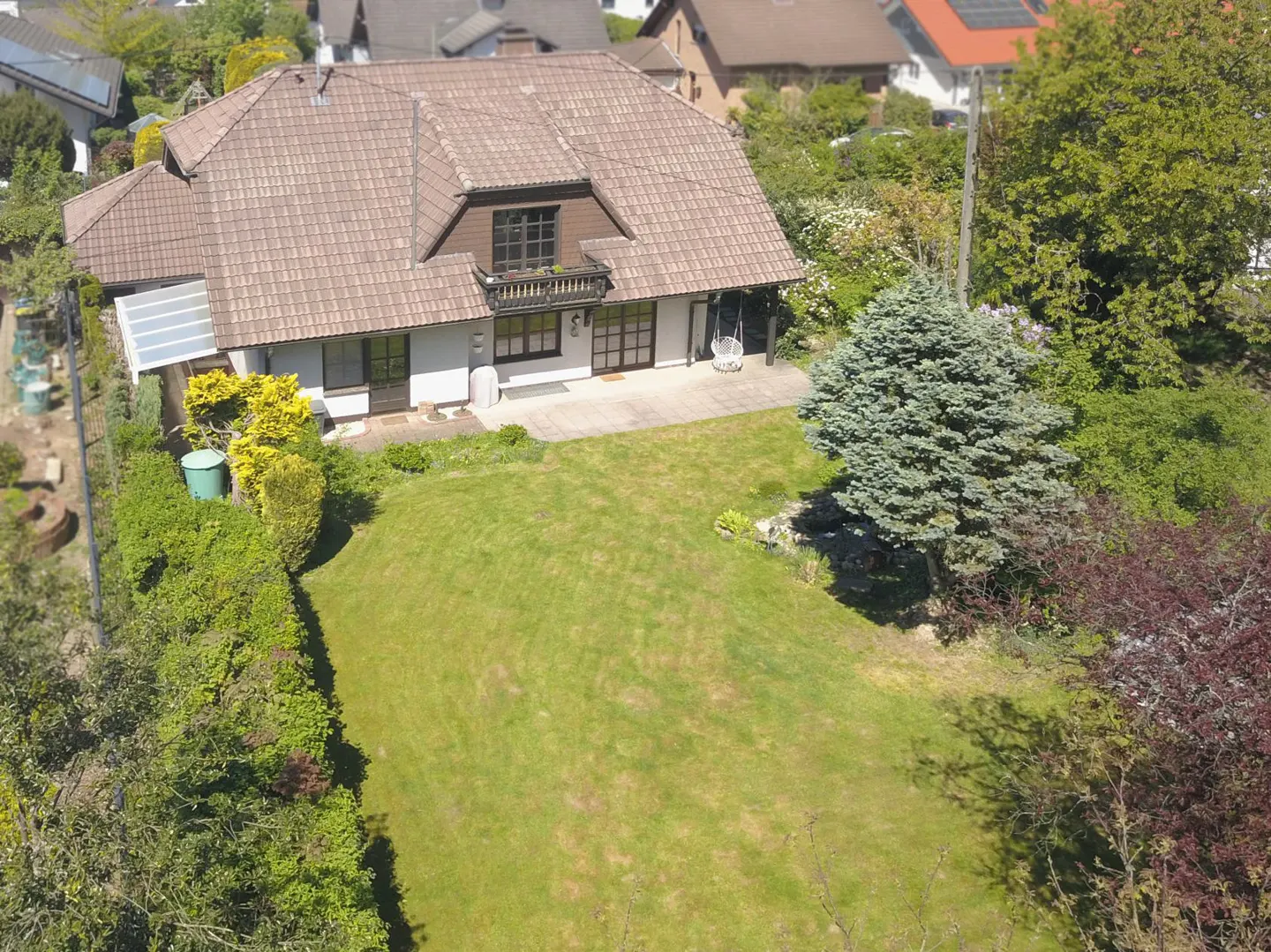 Aerial view of a white house with a brown roof, a balcony, and a large green lawn surrounded by trees and bushes.