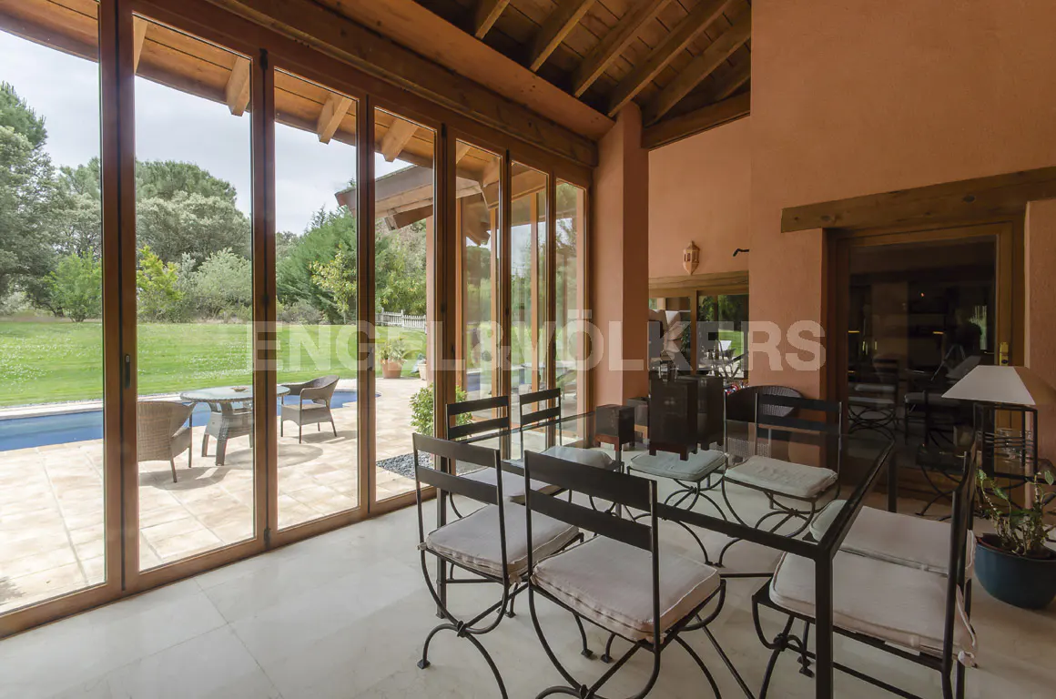 Indoor dining area with a glass table and black chairs, wooden ceiling, and large glass doors leading to a patio and green lawn.