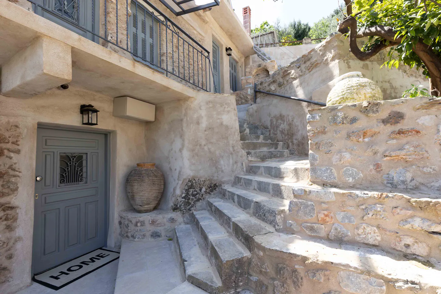 Exterior of a stone house with a gray door, stone steps, and a large clay pot. A "HOME" doormat sits in front of the door.
