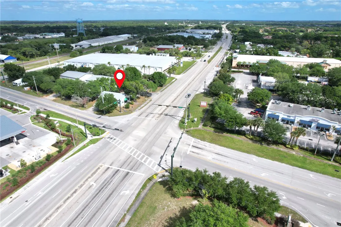 Aerial view of a property at a crossroads, marked with a red pin. Roads intersect, surrounded by trees and buildings under a blue sky.