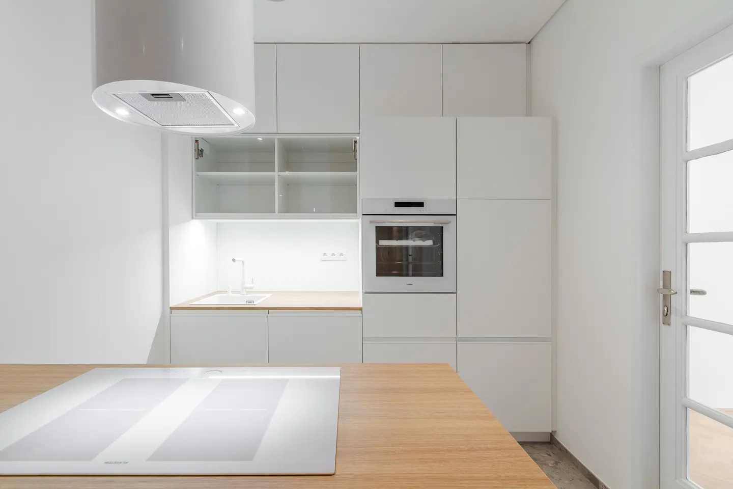 Bright, modern kitchen with white cabinets, oven, and range hood. A light wood island features a white cooktop. A white door is on the right.