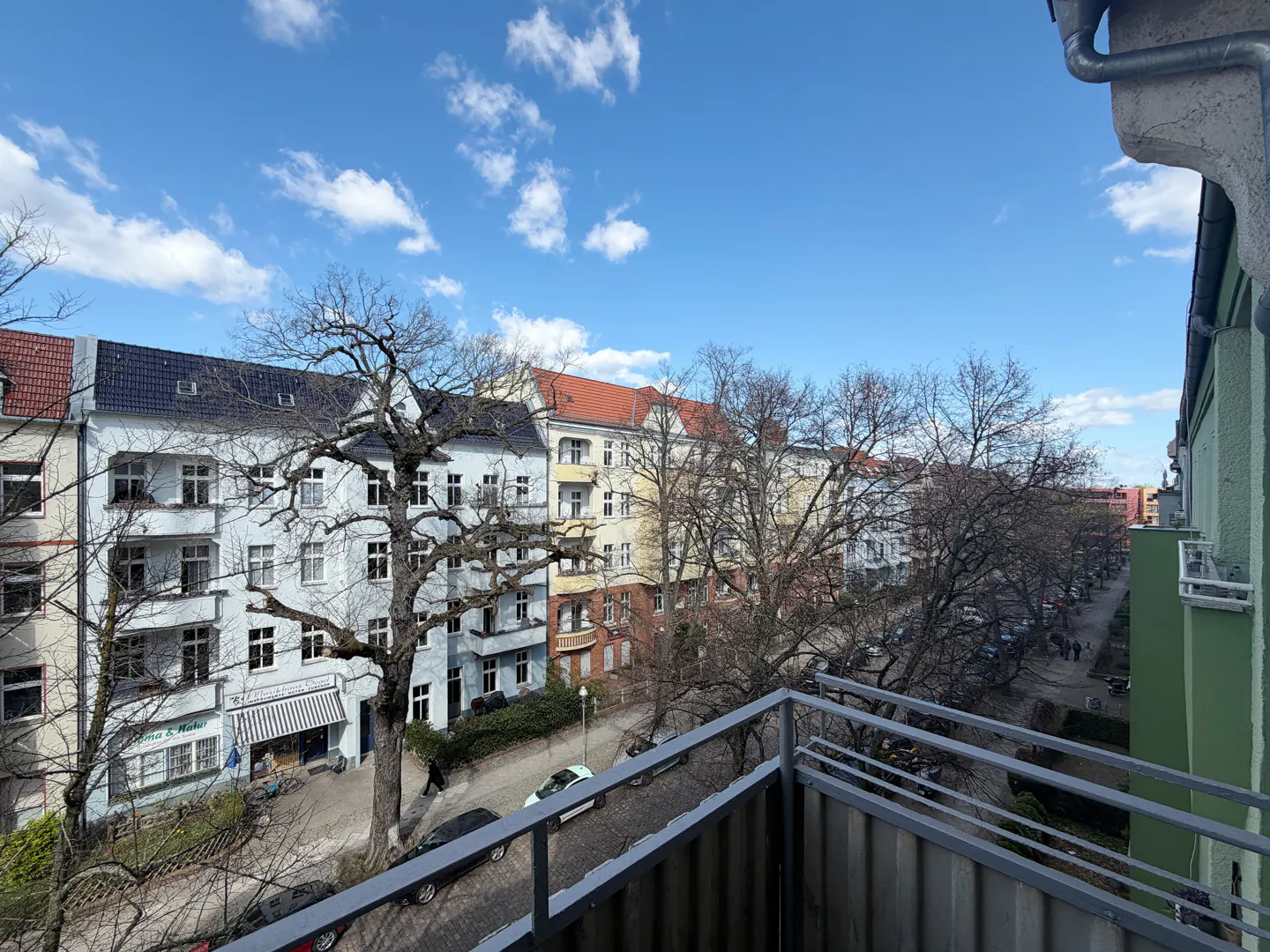 View from a balcony overlooking a street with colorful buildings and bare trees under a blue sky with clouds.