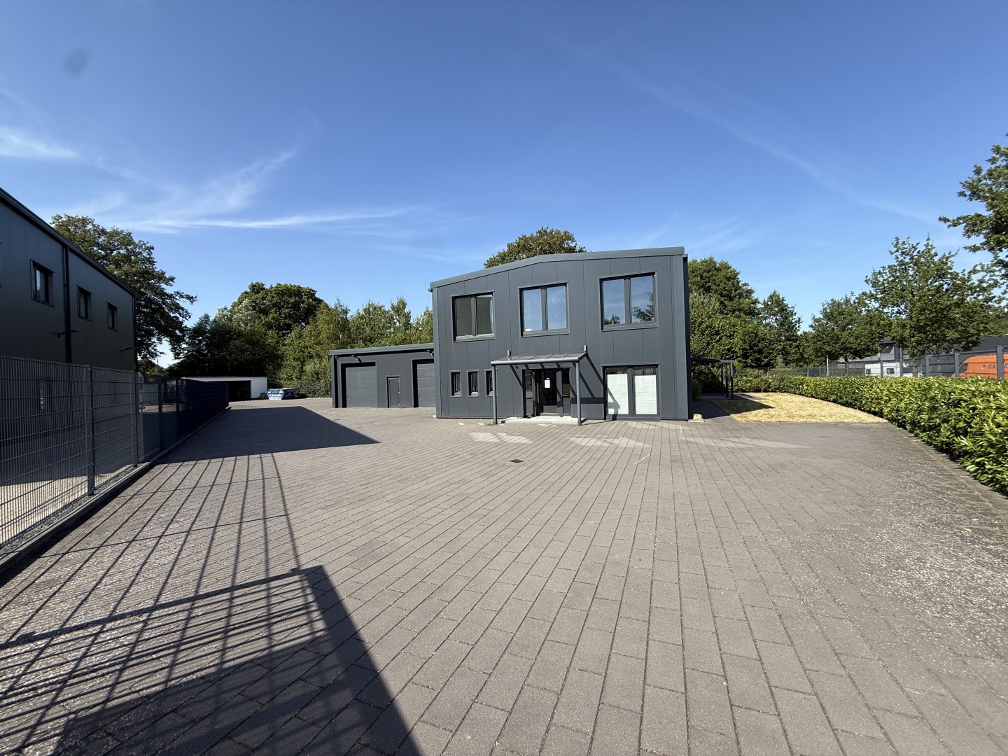 Modern dark gray commercial building with a paved parking lot under a clear blue sky, surrounded by trees and a fence. [AI-generated text]