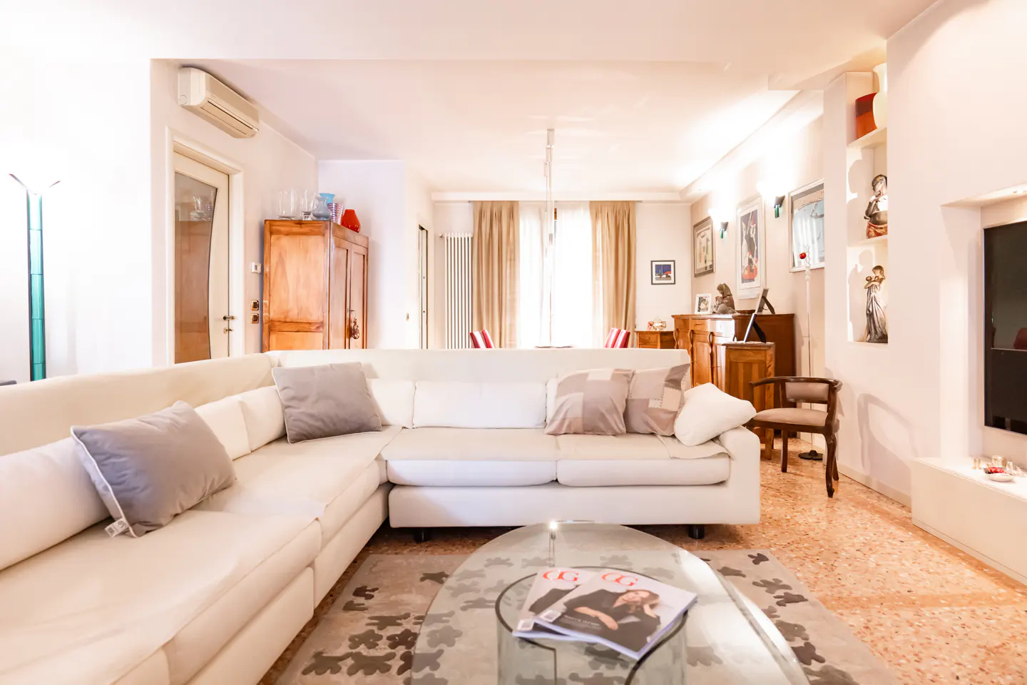 Bright living room with a large white sectional sofa, glass coffee table, and beige curtains in a sunlit, modern home.
