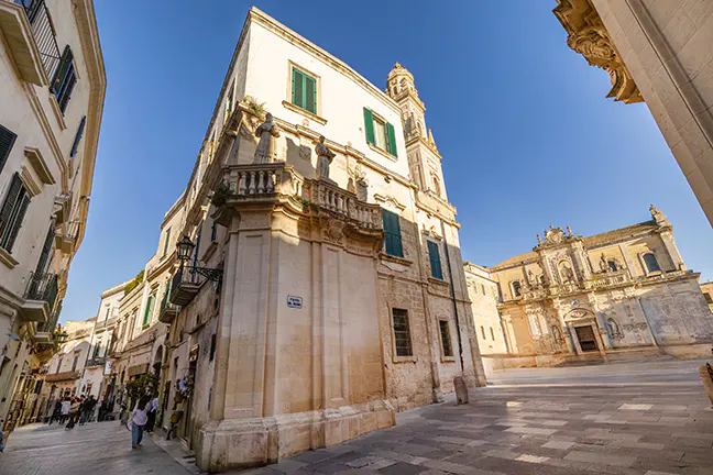 Low angle shot of Lecce, Italy, featuring cream-colored buildings with green shutters and a church in the background.