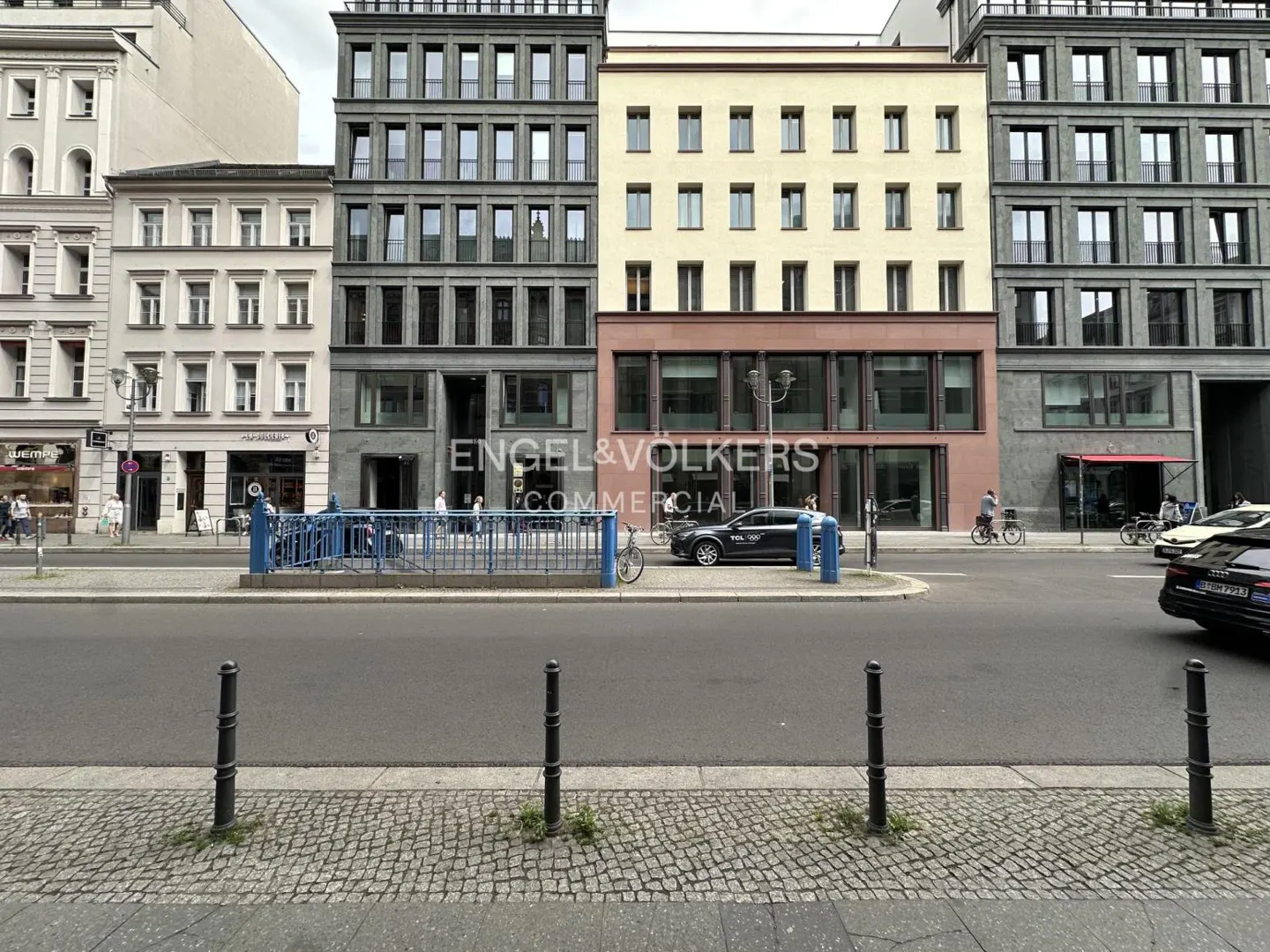 Street view of a city block with a mix of historic and modern buildings, a blue metal fence, and cars parked on the road.