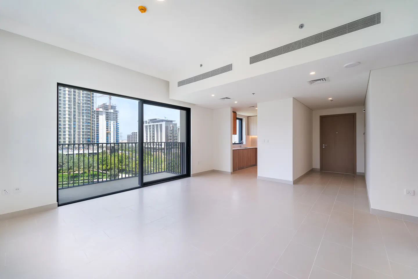 Bright, empty apartment with white walls and tile floors. A sliding glass door leads to a balcony with city views. A kitchen and hallway are visible.