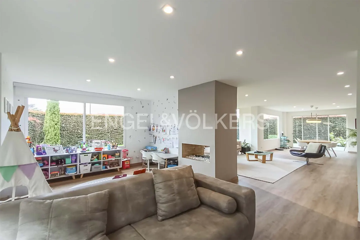 Spacious open-plan living area with a grey sofa, a central fireplace, a children's play corner, and a bright dining space.
