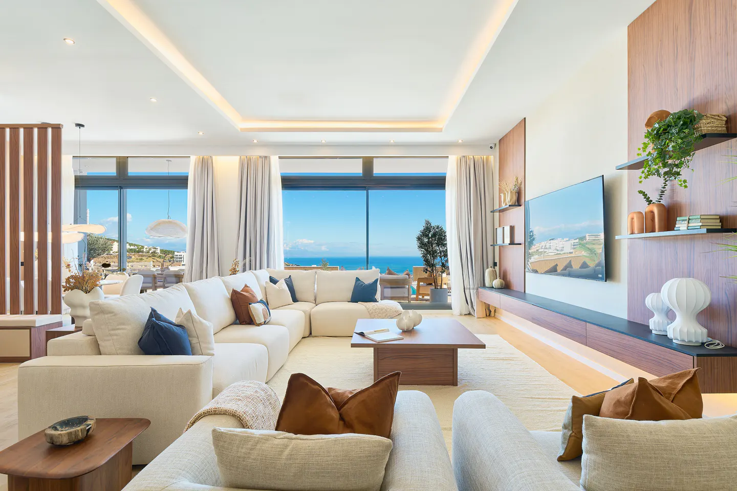 Bright living room with a white sectional sofa, wood accents, and a large window overlooking the ocean. A TV is mounted on the wall.