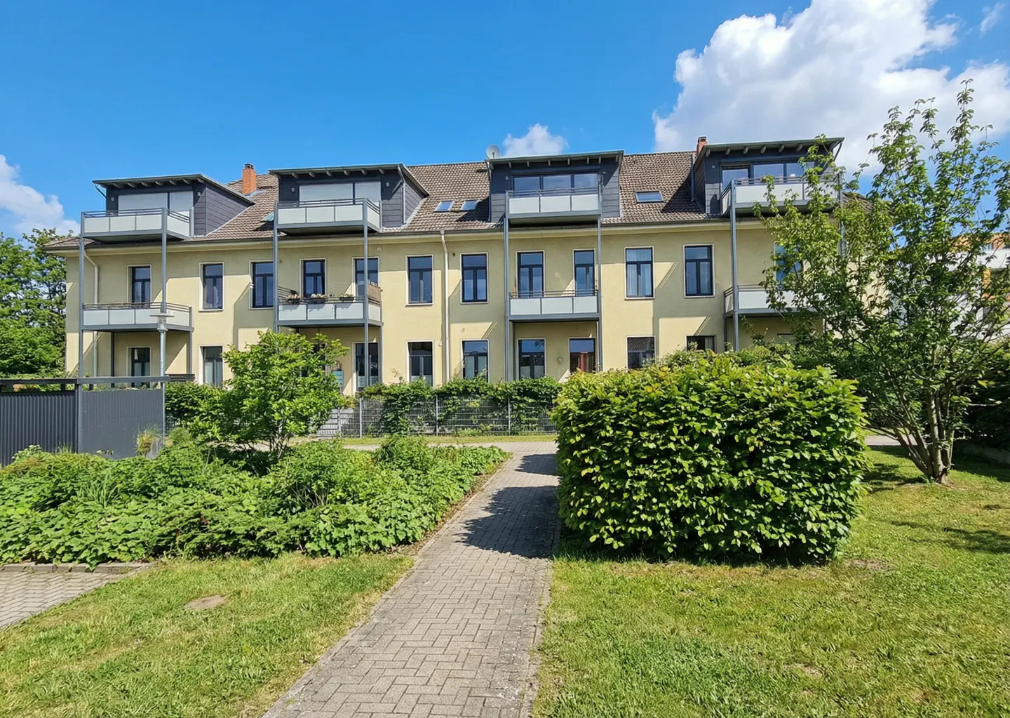 Three-story yellow apartment building with balconies, dark windows, and a brick walkway leading to the entrance. Green bushes and trees surround the building.