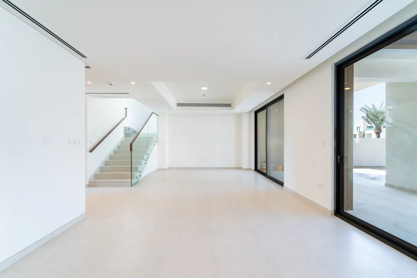 Bright, empty room with white walls, light tile floor, and a staircase with glass railing. Sliding glass doors lead to an outdoor patio.