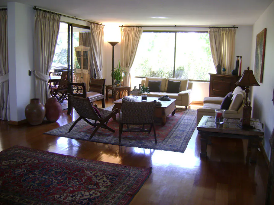 Bright living room with hardwood floors, beige sofas, and large windows with light curtains. A red and blue rug adds color.