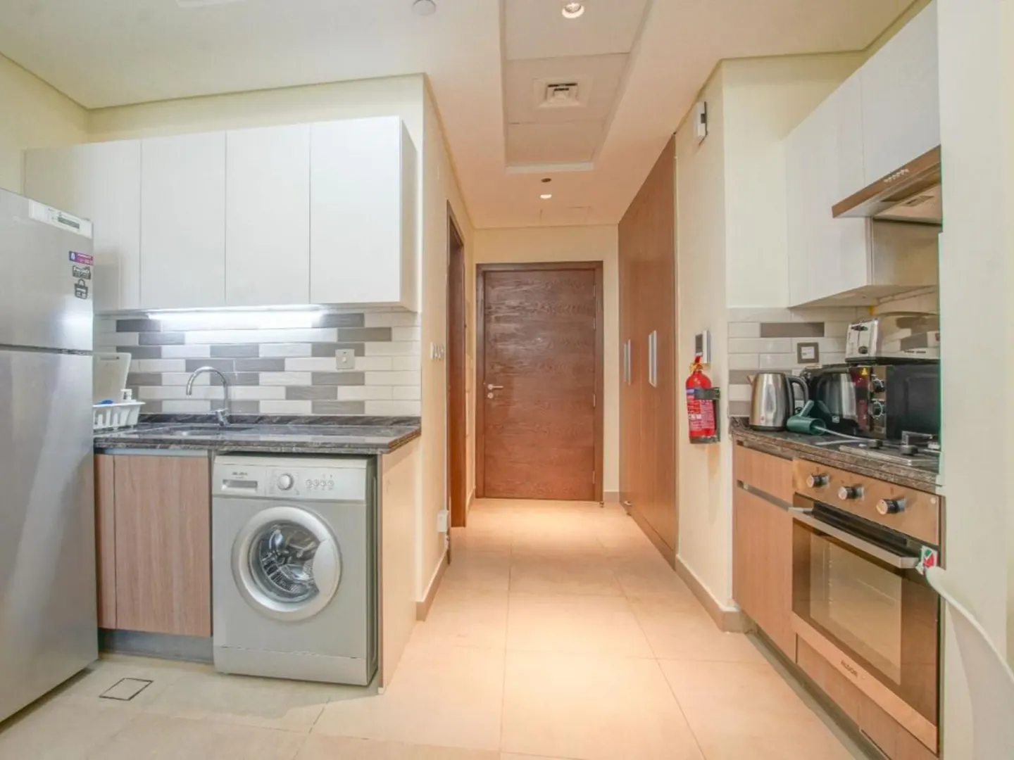 A modern kitchen with white cabinets, stainless steel appliances, and a washing machine. A hallway leads to a brown door.
