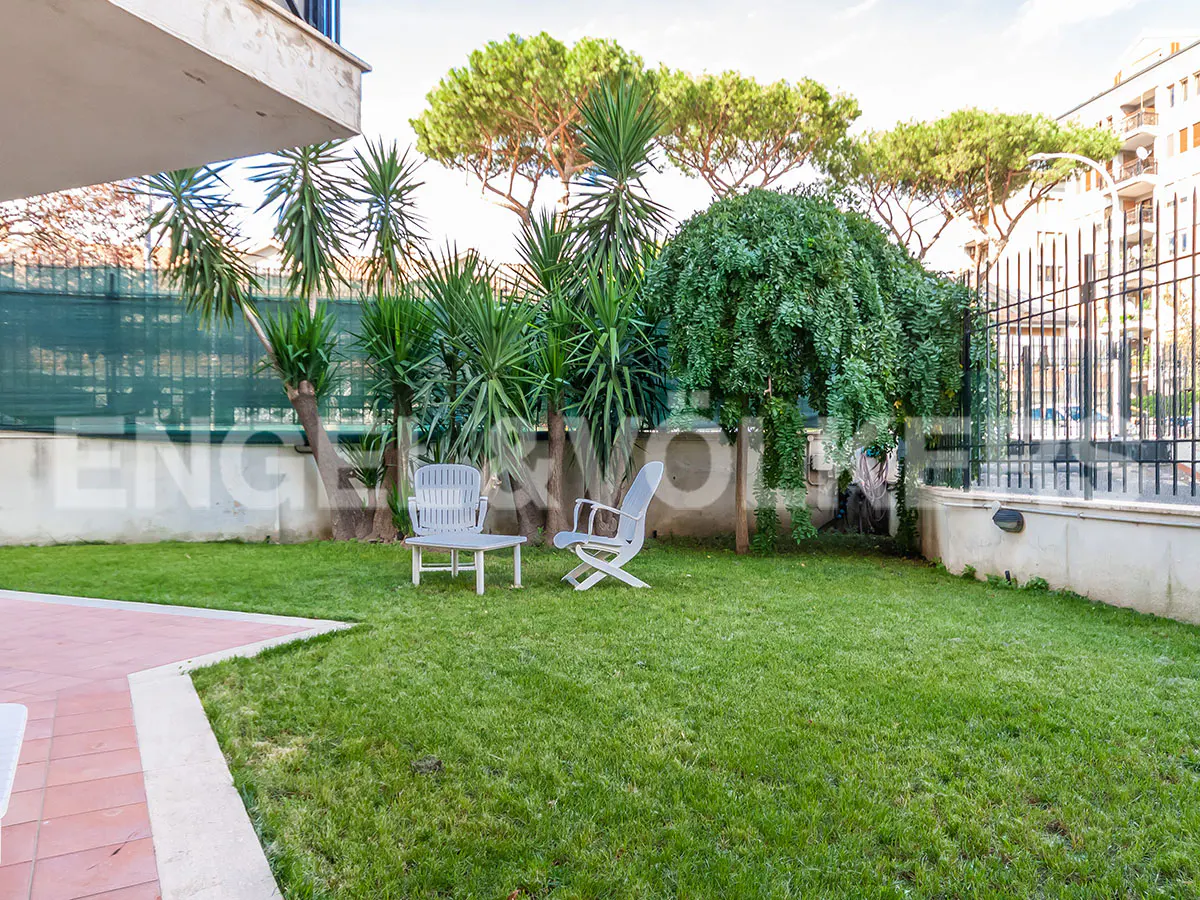 A green lawn with two white chairs, palm trees, and a building in the background.