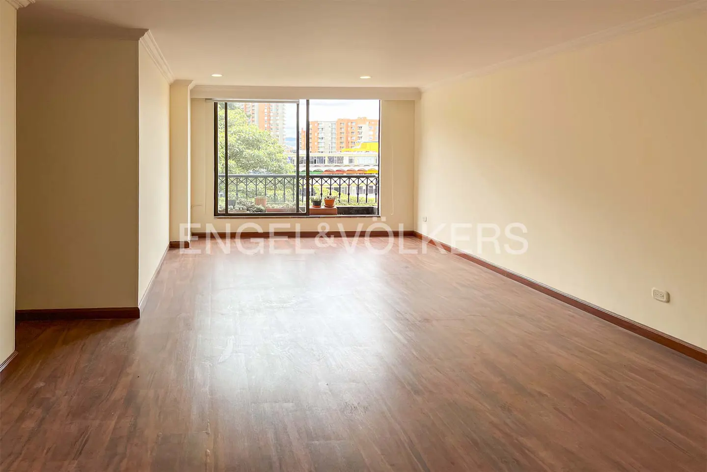 A spacious, empty living room with hardwood floors and a large window overlooking a city. Walls are light beige. Engel & Volkers logo is visible.
