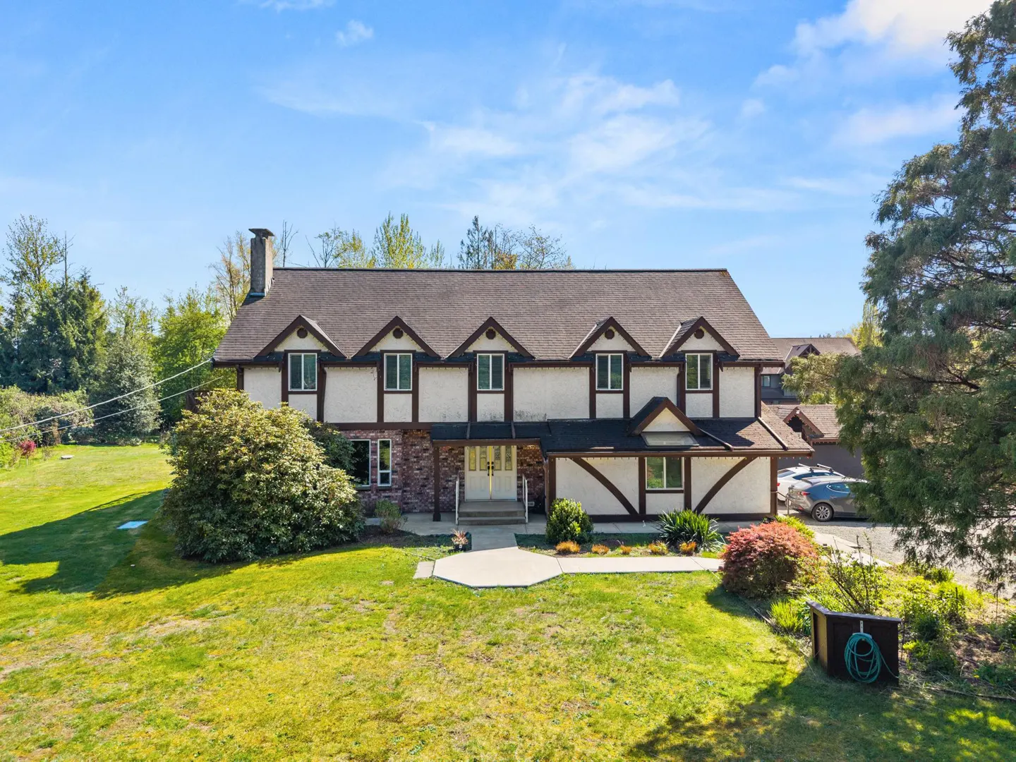 Two-story Tudor-style house with white stucco, brown trim, and a dark roof, surrounded by a green lawn and trees under a blue sky.